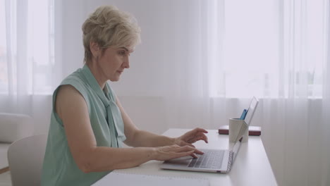 Una-Mujer-Anciana-Está-Charlando-En-Las-Redes-Sociales-A-Través-De-Internet-Usando-Una-Computadora-Portátil-Escribiendo-Mensajes-En-El-Teclado-Sentada-En-La-Mesa